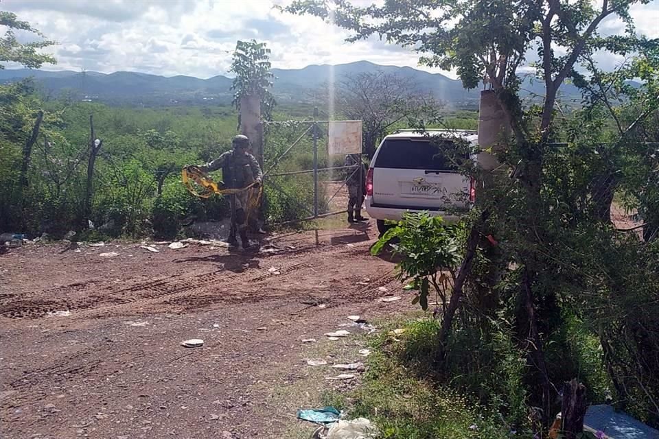 Una puerta con malla ciclónica sostenida por dos tubos de acero es custodiada por cuatro elementos del Ejército Mexicano y dos camionetas artilladas.