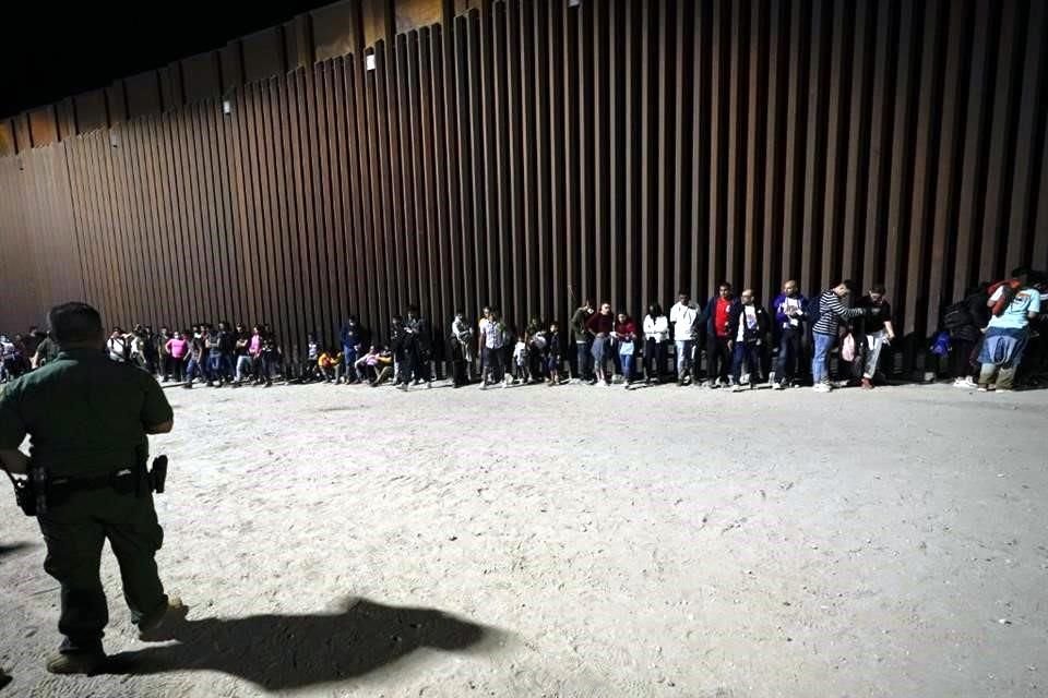 Migrantes esperan pegados al muro fronterizo en el área de Arizona mientras la Patrulla fronteriza de EU los procesa.