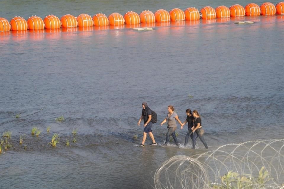 Migrantes caminan entre alambre de púas y las boyas naranjas en el Río Bravo.