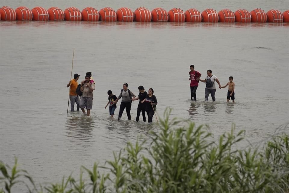 Migrantes que cruzaron el río Bravo desde México caminan frente a enormes boyas colocadas como una barrera fronteriza flotante en el río, el 12 de julio de 2023, en Eagles Pass, Texas.