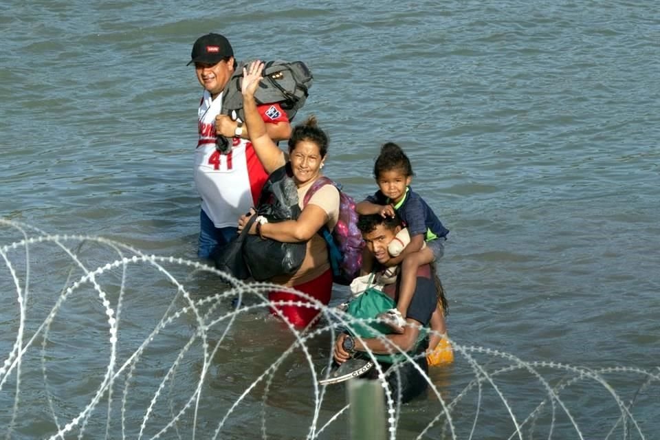 Migrantes saludan mientras caminan cerca de alambre de púas en el agua a lo largo de la frontera del Río Bravo.