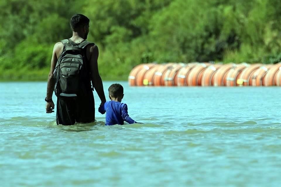 Migrantes pasan junto a grandes boyas que se utilizan como barrera fronteriza flotante en el Río Bravo.