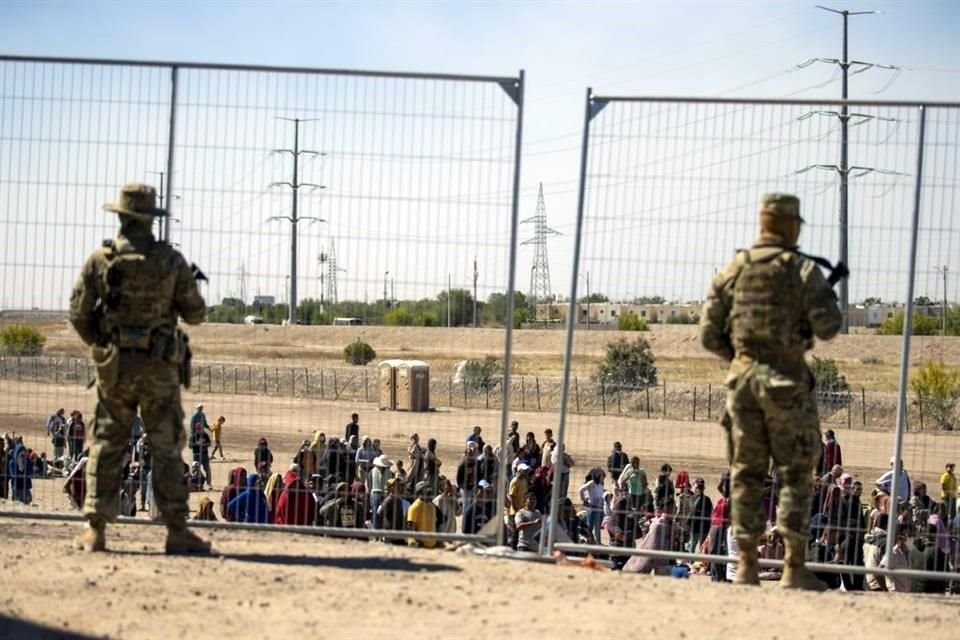 Los migrantes esperan en fila junto a la valla fronteriza bajo la vigilancia de la Guardia Nacional de Texas para ingresar a El Paso.