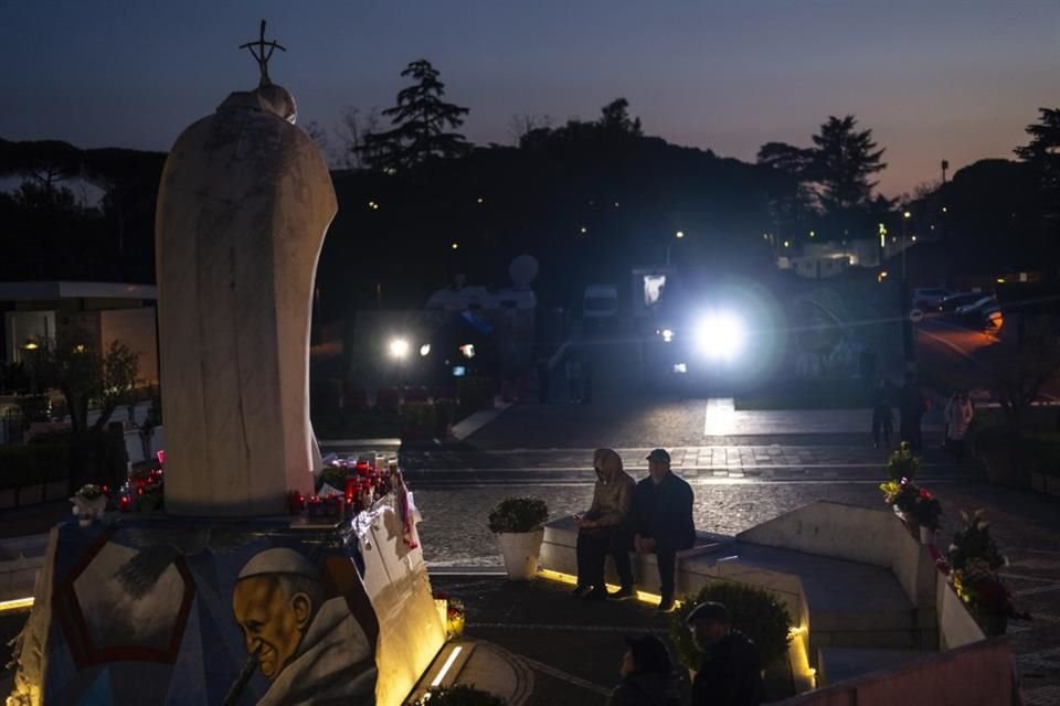 Personas sentadas frente a una estatua del Papa Juan Pablo II afuera del hospital donde el Papa Francisco está internado.